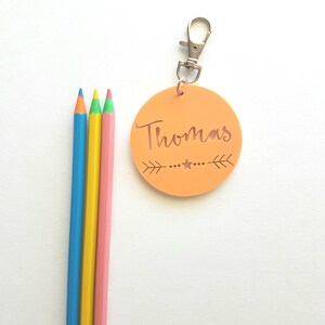 May include: A peach-colored round keychain with the name "Thomas" written in a cursive font. The keychain has a silver clip and a small star design. Three colored pencils are in the background.