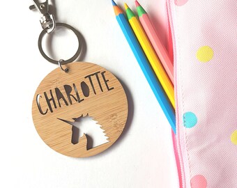 Puede incluir: Un llavero de madera con el nombre "CHARLOTTE" grabado en él. El llavero presenta un recorte de una cabeza de unicornio. El llavero está unido a un llavero plateado. El llavero está al lado de un estuche de lápices rosa con lunares blancos y tres lápices de colores.