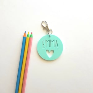 May include: A light teal keychain with the name "EMMA" cut out, along with a heart shape. The keychain has a silver clasp. Three colored pencils in blue, yellow, and pink are next to the keychain.