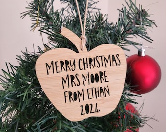 May include: A wooden apple-shaped Christmas ornament with the text "Merry Christmas Mrs. Moore From Ethan 2022" hanging on a Christmas tree.
