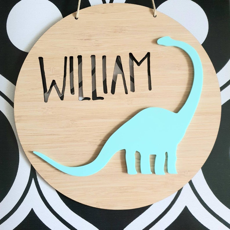 May include: A round wooden sign with the name "WILLIAM" cut out in the center. A light blue dinosaur silhouette is attached to the sign.