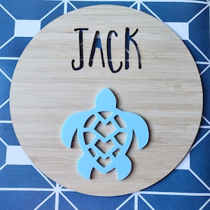 以下が含まれることがあります： 薄い木製の丸い看板に、黒い文字で「JACK」という名前が切り抜かれています。看板の中央には、薄い青色のカメの切り抜きがあります。