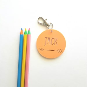 May include: A round, orange name tag with a silver keychain. The name tag has the name "JACK" cut out of the center and a small arrow design on either side of the name. Three colored pencils are in the background.