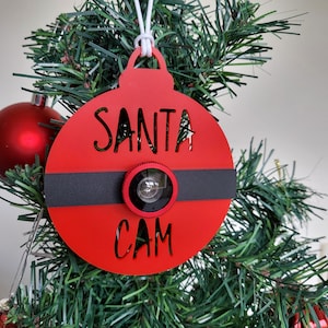 May include: Red Christmas ornament with a black band and a small camera lens in the center. The ornament says "Santa Cam" in black lettering.