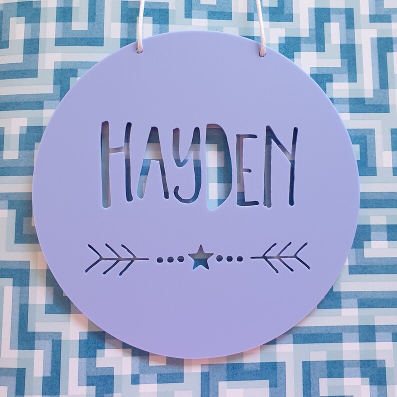 May include: A light purple round wooden sign with the name Hayden cut out in the center. The sign has a white string for hanging and a small star and arrow design at the bottom.