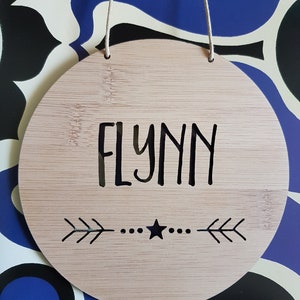 May include: A round, light brown wooden sign with the name "FLYNN" written in black lettering. The sign has a black star and two arrows on either side of the star.