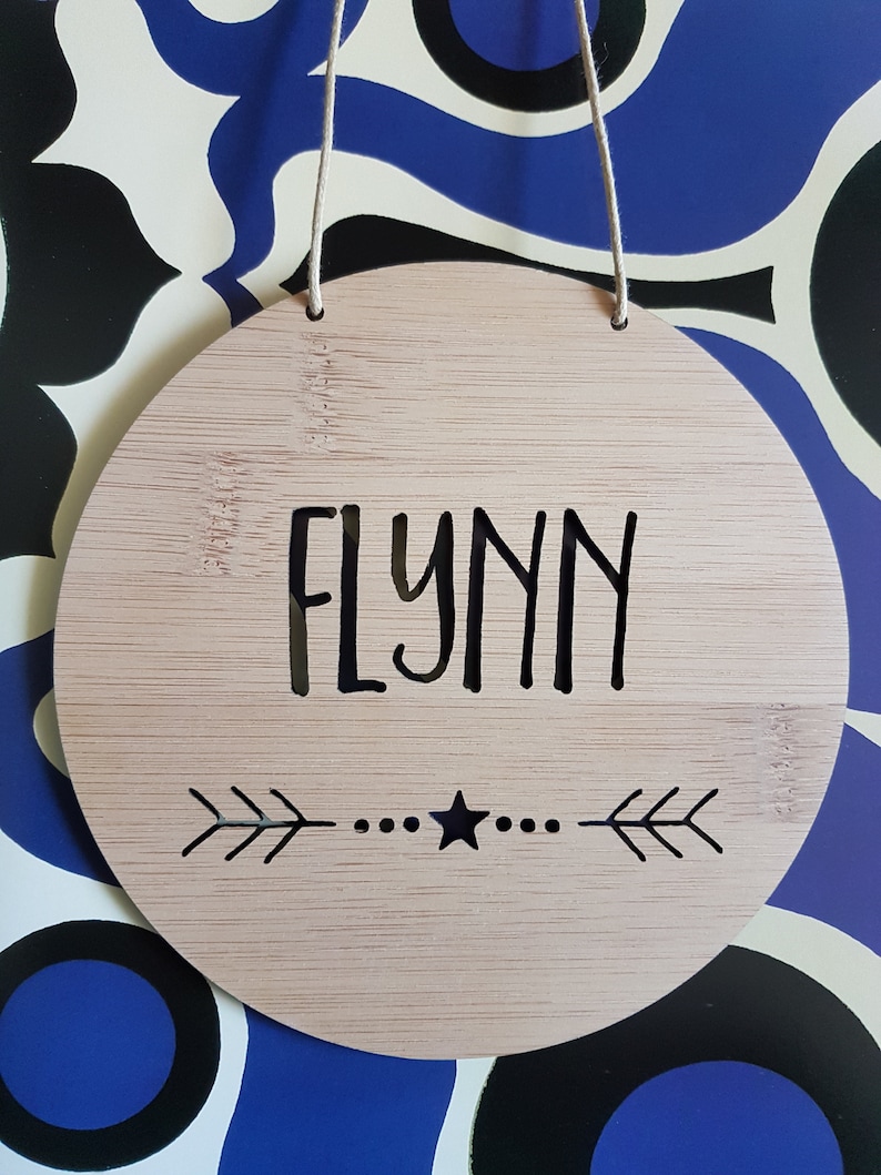 May include: A round, light brown wooden sign with the name "FLYNN" written in black lettering. The sign has a black star and two arrows on either side of the star.