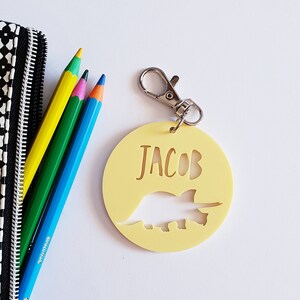 May include: A yellow round keychain with a dinosaur silhouette and the name "JACOB" written in gold lettering. The keychain has a silver metal clasp.