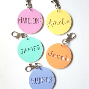 May include: Five colorful round keychains with names written on them. The keychains are pink, yellow, light green, orange, and light purple. The names are Madeleine, Amelia, James, Jacob, and Hudson.