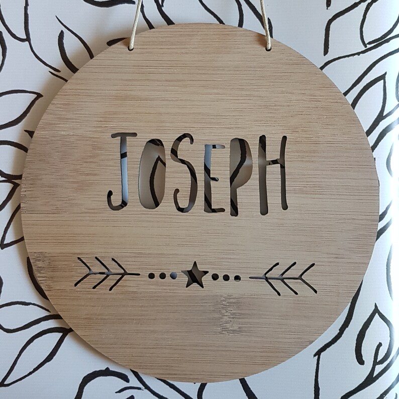 May include: A round wooden sign with the name "Joseph" cut out in the center. The sign has a natural wood finish and features a small star and two arrows on the bottom.