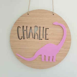 May include: A round wooden sign with the name "CHARLIE" cut out in the center. A pink dinosaur silhouette is attached to the right side of the sign.