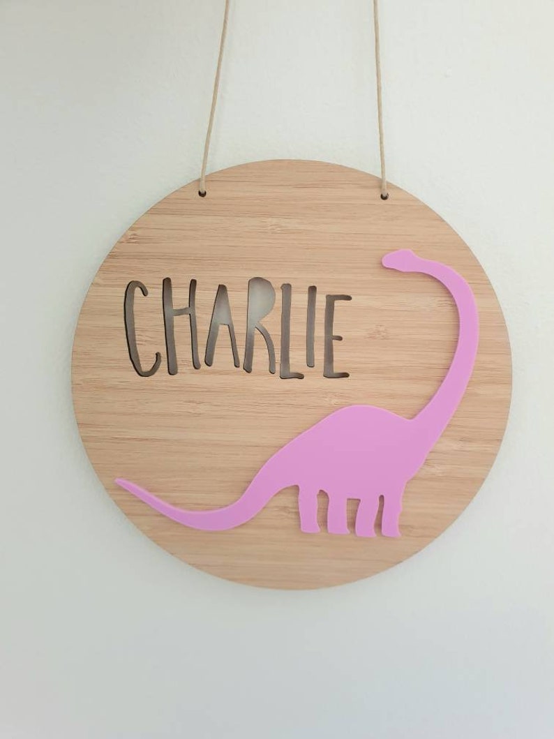 May include: A round wooden sign with the name "CHARLIE" cut out in the center. A pink dinosaur silhouette is attached to the right side of the sign.