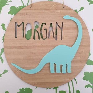 May include: A round wooden sign with the name "MORGAN" cut out in the center. A light blue dinosaur silhouette is on the sign.