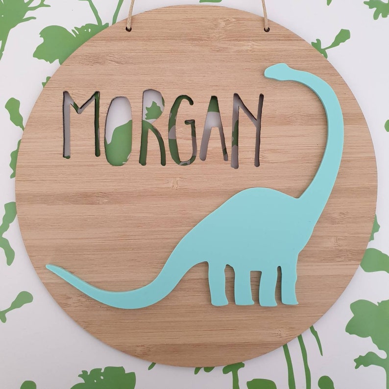 May include: A round wooden sign with the name "MORGAN" cut out in the center. A light blue dinosaur silhouette is on the sign.