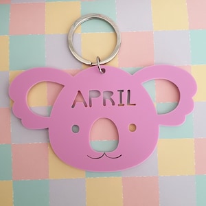May include: A pink koala-shaped keychain with the month "APRIL" cut out. The keychain has a silver metal ring for attaching keys. The koala's face features cut-out eyes and a smiling mouth. The background is a pastel checkered pattern.