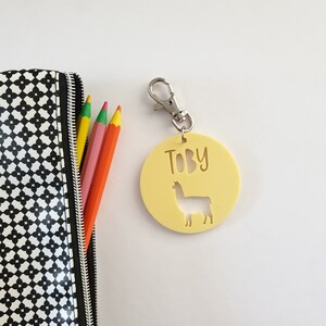 May include: A yellow keychain with the name "Toby" and a llama cutout. The keychain is attached to a silver clasp. A black and white patterned pencil case and colored pencils are in the background.