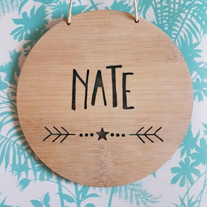 May include: A round wooden sign with the name "NATE" written in black lettering. The sign has a star and arrows carved into the wood.