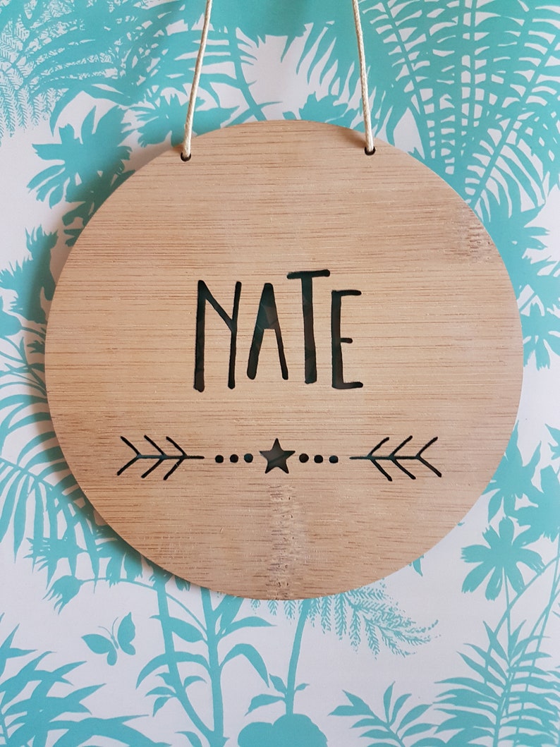 May include: A round wooden sign with the name "NATE" written in black lettering. The sign has a star and arrows carved into the wood.