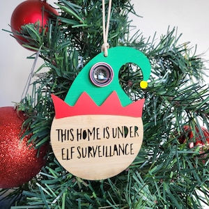 May include: A green and red elf ornament with a camera lens and the text "This home is under elf surveillance".
