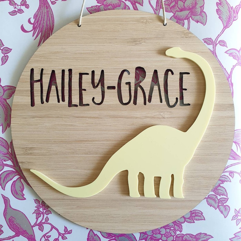 May include: A round wooden sign with the name "HAILEY-GRACE" cut out in the center. A yellow dinosaur silhouette is attached to the sign.