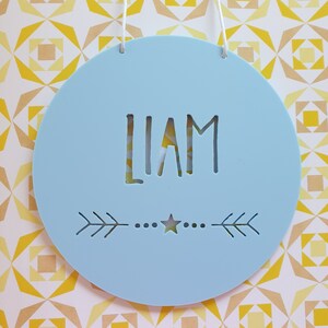 May include: A light blue round wooden sign with the name "LIAM" cut out in the center. The sign has a small star and two arrows on the bottom.