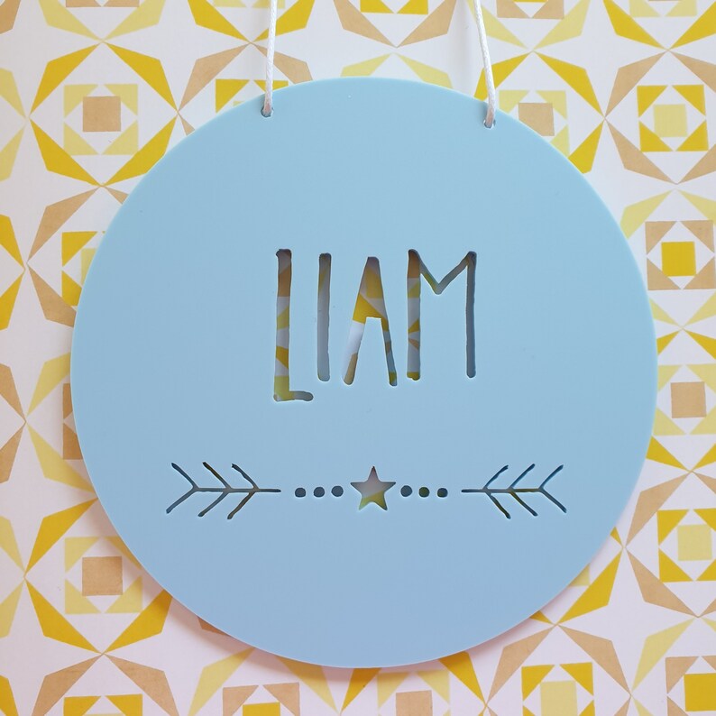 May include: A light blue round wooden sign with the name "LIAM" cut out in the center. The sign has a small star and two arrows on the bottom.