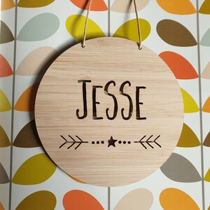 May include: A round wooden sign with the name "Jesse" engraved in a cursive font. The sign has a small star and two arrows on either side of the name.