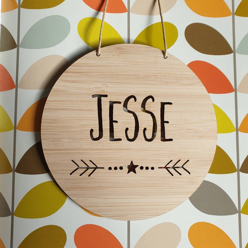 May include: A round wooden sign with the name "Jesse" engraved in a cursive font. The sign has a small star and two arrows on either side of the name.
