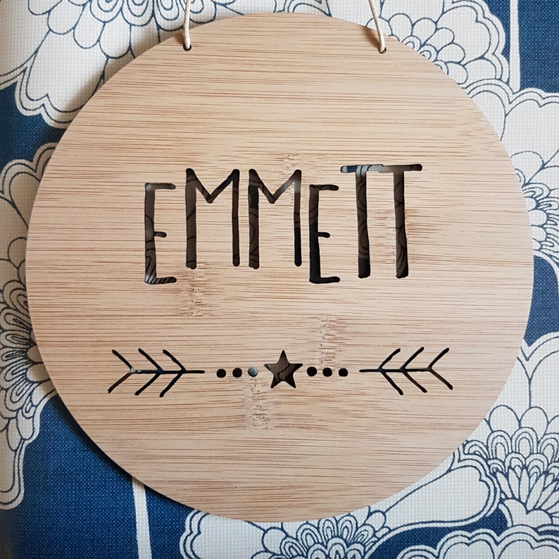 May include: A round, wooden sign with the name "EMMETT" cut out in the center. The sign has a small star and arrow design below the name.