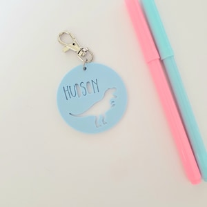 May include: Light blue round keychain with a dinosaur silhouette and the name "HUDSON" in white lettering. The keychain has a silver metal clasp. Two pink and blue markers are in the background.
