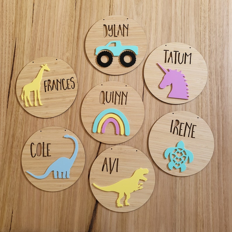 May include: Seven wooden round signs with names written on them. Each sign has a different colored cutout design. The designs include a monster truck, a unicorn, a rainbow, a dinosaur, a giraffe, a turtle, and a dinosaur.