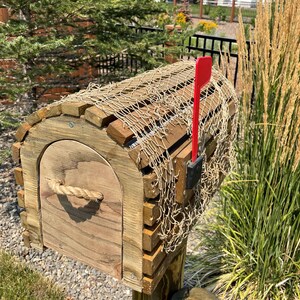 May include: A wooden mailbox with a curved roof and a rope handle. The mailbox is decorated with a fishing net and has a red flag. The mailbox is mounted on a wooden post and is surrounded by greenery. The mailbox is made of wood.