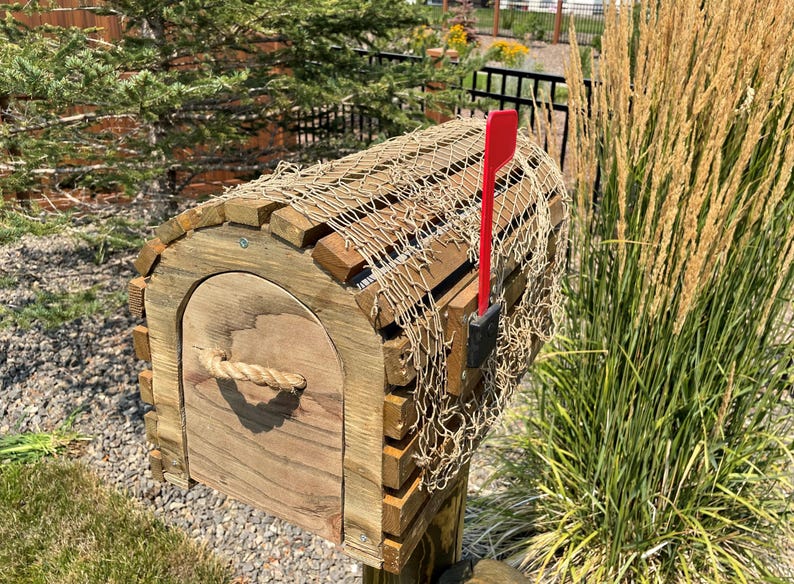May include: A wooden mailbox with a curved roof and a rope handle. The mailbox is decorated with a fishing net and has a red flag. The mailbox is mounted on a wooden post and is surrounded by greenery. The mailbox is made of wood.