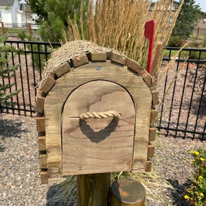 May include: A wooden mailbox with a rope handle and a red flag. The mailbox is made from weathered wood and has a thatched roof. It is mounted on a wooden post and surrounded by gravel.