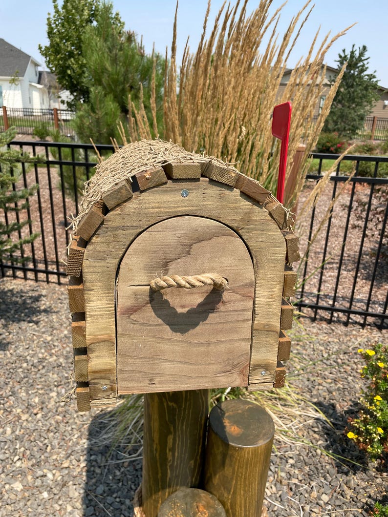 May include: A wooden mailbox with a rope handle and a red flag. The mailbox is made from weathered wood and has a thatched roof. It is mounted on a wooden post and surrounded by gravel.