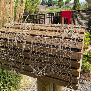 May include: A wooden mailbox with a decorative fishing net draped over the top. The mailbox has a red flag and is mounted on a wooden post. The mailbox is surrounded by greenery and yellow flowers.