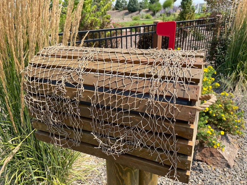 May include: A wooden mailbox with a decorative fishing net draped over the top. The mailbox has a red flag and is mounted on a wooden post. The mailbox is surrounded by greenery and yellow flowers.
