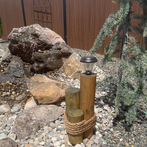 May include: A solar-powered garden light fixture made of wood posts and a clear plastic lamp. The fixture is wrapped with rope and sits on a bed of small stones. A small waterfall and greenery are in the background.