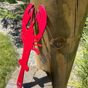 May include: A vibrant red metal lobster-shaped bottle opener. The opener is a silhouette of a lobster with a handle and a small hole at the end. The lobster is displayed outdoors, leaning against a wooden post.