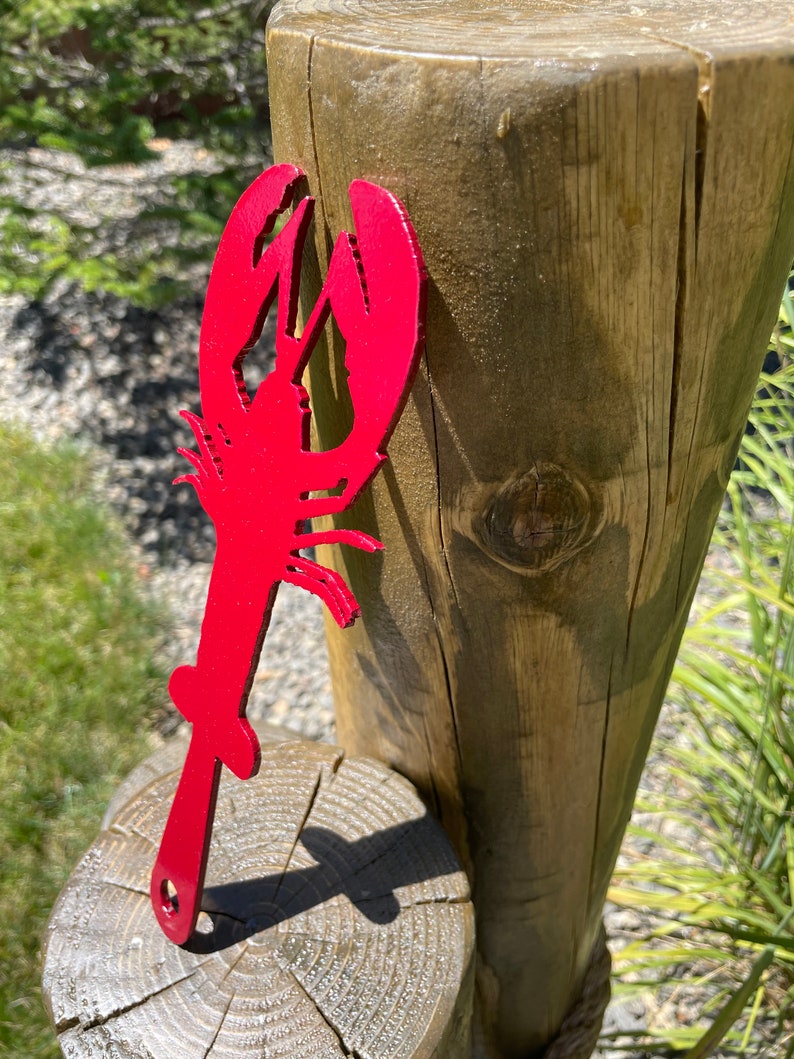May include: A vibrant red metal lobster-shaped bottle opener. The opener is a silhouette of a lobster with a handle and a small hole at the end. The lobster is displayed outdoors, leaning against a wooden post.