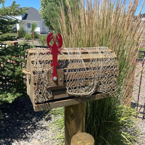 May include: A wooden mailbox designed to look like a lobster trap, complete with a fishing net. It features a red metal lobster decoration. The mailbox is mounted on a wooden post, evoking a seaside aesthetic. The overall design is reminiscent of a coastal theme.