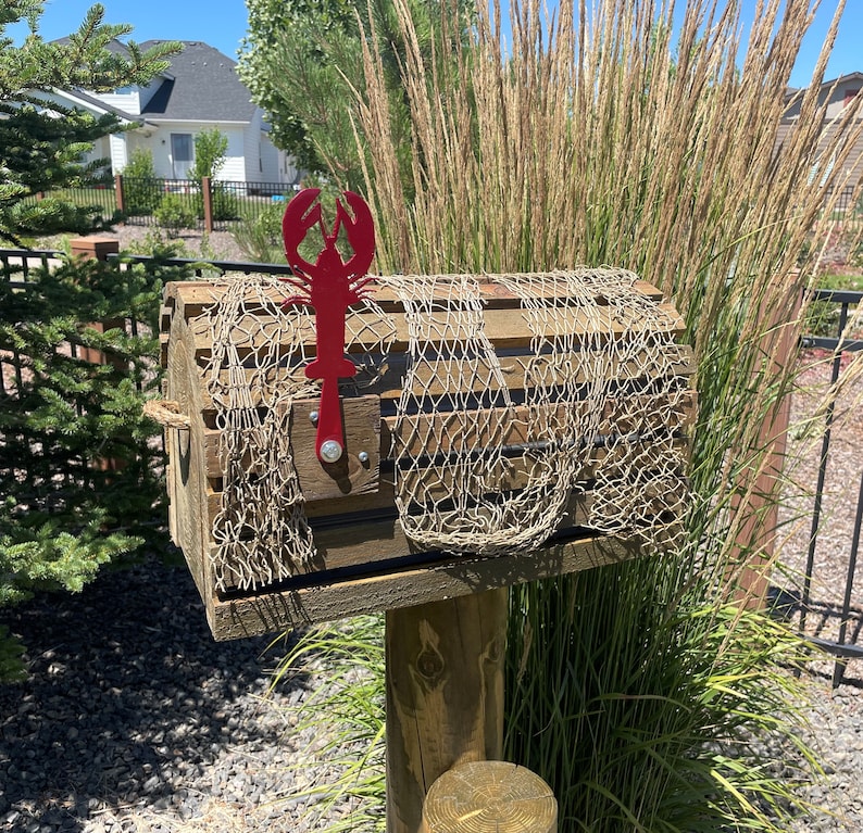 May include: A wooden mailbox designed to look like a lobster trap, complete with a fishing net. It features a red metal lobster decoration. The mailbox is mounted on a wooden post, evoking a seaside aesthetic. The overall design is reminiscent of a coastal theme.