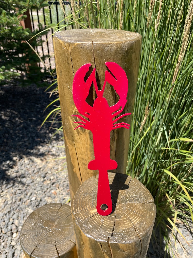 May include: A vibrant red metal lobster garden stake. The lobster features detailed claws and body segments, with a handle and a hole for insertion. The stake is displayed outdoors, likely in a garden.