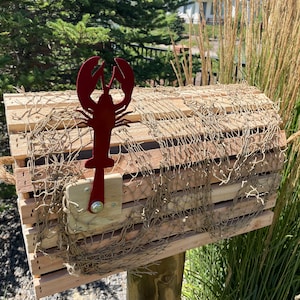 May include: A wooden lobster trap-style mailbox with a red metal lobster silhouette. The mailbox is covered with netting and mounted on a wooden post. The design has a coastal or nautical theme, with a rustic aesthetic.