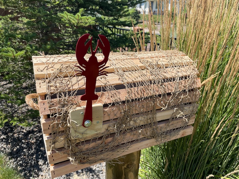 May include: A wooden lobster trap-style mailbox with a red metal lobster silhouette. The mailbox is covered with netting and mounted on a wooden post. The design has a coastal or nautical theme, with a rustic aesthetic.