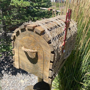 May include: A weathered wooden mailbox with a curved front door and rope handle. The mailbox is covered with a decorative net and has a red metal flag. The design is rustic, with a natural wood finish. The mailbox is set against a backdrop of greenery.