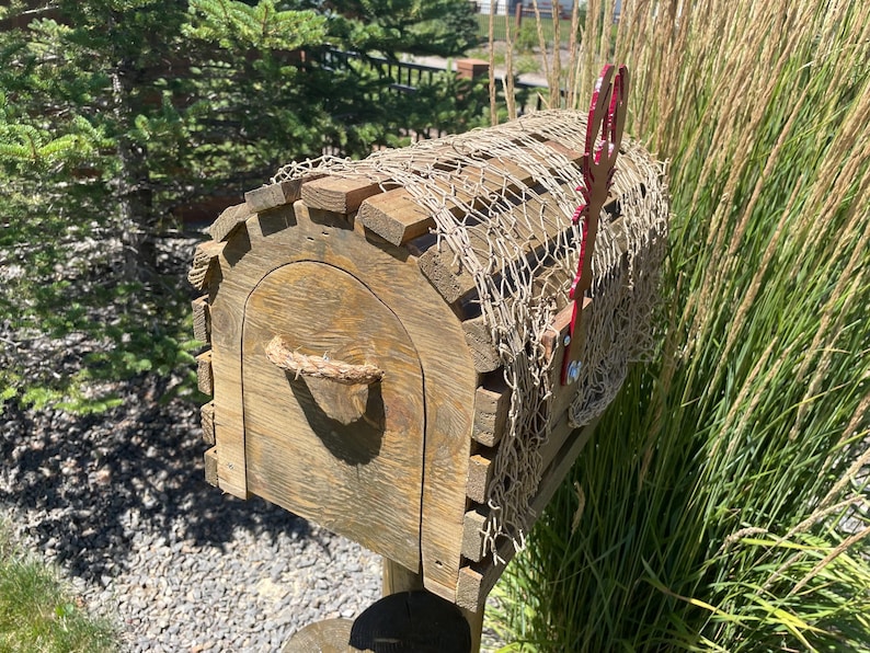 May include: A weathered wooden mailbox with a curved front door and rope handle. The mailbox is covered with a decorative net and has a red metal flag. The design is rustic, with a natural wood finish. The mailbox is set against a backdrop of greenery.