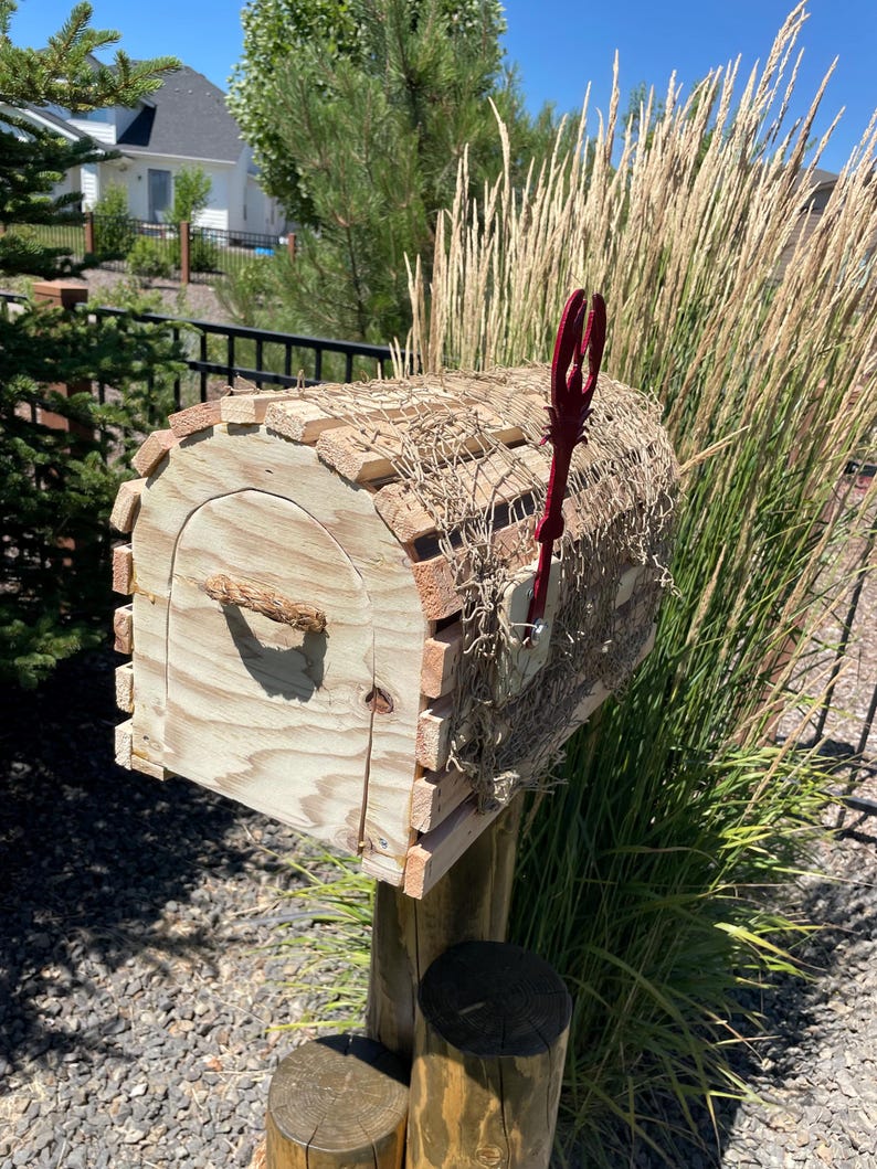 May include: A wooden mailbox with a rustic design. The mailbox is made of light-coloured wood with a curved top and a red trident-shaped flag. The mailbox is mounted on a wooden post and surrounded by tall grass.