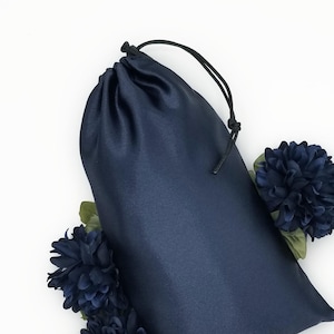 May include: A navy blue satin drawstring bag with a black cord closure. The bag is surrounded by three navy blue artificial flowers with green leaves, set against a white background. The bag appears to be empty.