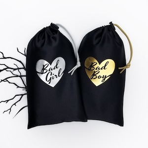 May include: Two black satin drawstring bags with silver and gold foil hearts. The bag on the left has a silver heart with the text "Bad Girl" and the bag on the right has a gold heart with the text "Bad Boy".